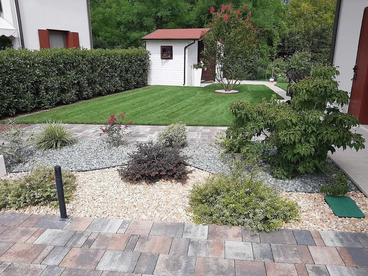 Backyard garden with paved patio, gravel area with shrubs, manicured lawn, white shed with red roof, and surrounding hedges and trees