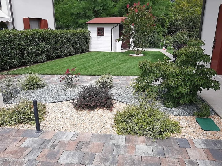 Backyard garden with paved patio, gravel area with shrubs, manicured lawn, white shed with red roof, and surrounding hedges and trees