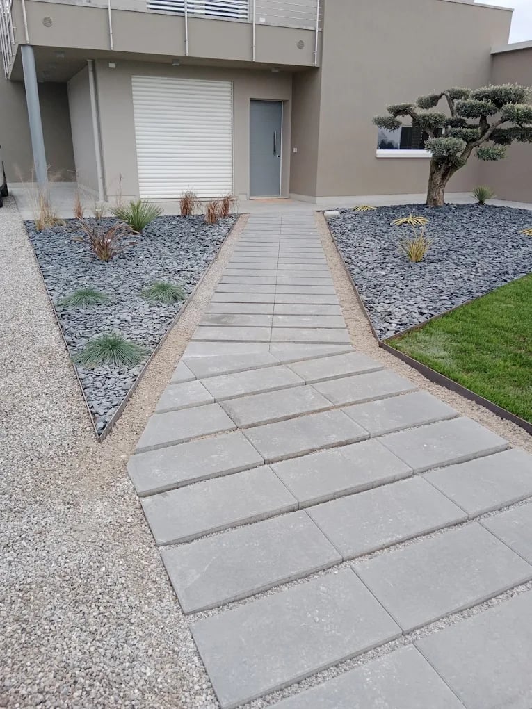 Modern house entrance with concrete paver walkway bordered by decorative gravel, small plants, and a topiary tree