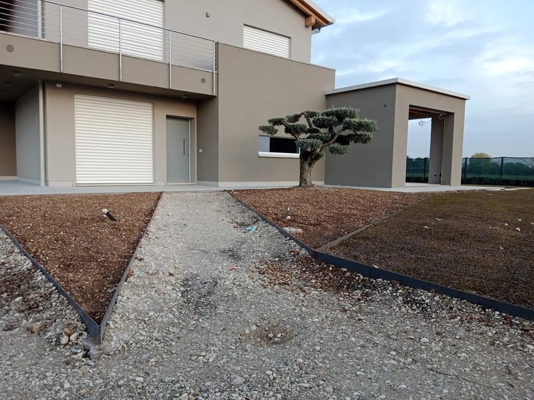 Modern beige house with gravel driveway, metal railings, and a small tree in landscaped yard under cloudy sky
